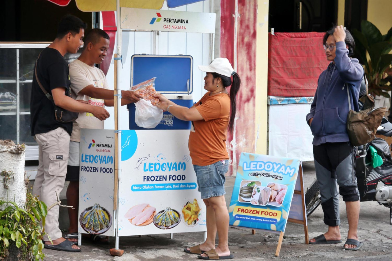 Fahmi didampingi terapis, Arbai, melayani pembeli di lapak usaha “Ledoyam” di Kota Malang, pada Jumat (17/10/2025). Lapak ini merupakan bantuan CSR PGN untuk mendukung program pelatihan wirausaha anak-anak autis. (Foto: Ali Masduki/jatimnow.com)